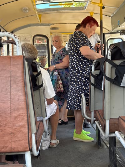 The image depicts the interior of a public transport bus, likely a trolleybus, with a sign visible that reads "46 РУБ." (46 rubles) indicating a fare. The bus appears to be in motion or stopped at a station, with several passengers visible.
On the right, a woman with short red hair is wearing a dark blue dress with white and yellow polka dots. She is holding a black object with a silver tip, possibly a cane or a device, in her right hand. She is facing forward, looking down.
To her left, a woman with blonde hair is wearing a floral print dress. She is looking to her right and holding a bag with a floral pattern.
A man with gray hair is seated on the left, facing away from the camera. He is wearing a white shirt and white shorts.
The bus interior features tan-colored seating with metal frames and a textured gray floor. Overhead, there are yellow handrails and lights. The scene suggests a typical public transportation environment in Russia, given the Cyrillic text and currency. The presence of a fare price sign for "46 rubles" suggests the location is in the Russian city of Shakhty, where such fares would be relevant.