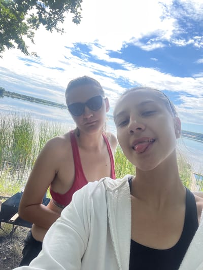 The image shows two young women in an outdoor setting. One woman in the foreground is wearing a red bikini top and sunglasses. She is leaning forward, with her arm resting on something dark that appears to be a grill. The other woman is behind her, leaning her head towards the camera and sticking out her tongue. She is wearing a black tank top and a white hoodie.
The scene appears to be near a body of water, possibly a lake or river, with tall grass and trees in the background. The sky is blue with white clouds, suggesting it is daytime and the weather is pleasant. The overall mood of the image seems playful and relaxed. There is no visible text in the image.