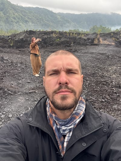 A man with a beard and short hair is taking a selfie in the foreground. He is wearing a black jacket and a checkered scarf. In the background, a woman with red hair is taking a photo with her phone. She is wearing a brown jacket and loose-fitting tan pants. The two individuals are standing on a rocky, dark-colored terrain that appears to be volcanic ash or lava rock. Behind them, lush green mountains rise up, partially shrouded in mist. The scene is set outdoors in a mountainous, potentially volcanic, landscape in Kintamani, Indonesia. The sky is overcast.