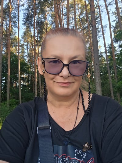This is a close-up, selfie-style, vertical photograph of a woman in an outdoor setting. The woman is wearing a black t-shirt and sunglasses with a beaded chain. She has blonde hair pulled back and is smiling gently. A black necklace with a clover charm and a bejeweled insect-shaped brooch are visible on her shirt. Behind her, a dense forest of pine trees is visible, with the sky peeking through the branches. The lighting suggests it is daytime, possibly late afternoon, given the warm tones and dappled sunlight. The scene evokes a sense of casual leisure and enjoyment of nature. The location is Daugavpils, Latvia. No text is visible in the image.
