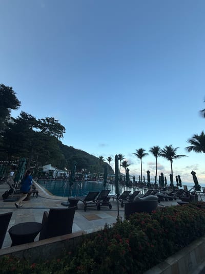 This media file captures a serene outdoor scene at what appears to be a resort or hotel in a tropical or sub-tropical setting, consistent with Rio de Janeiro, Brazil.
In the foreground, a person wearing a blue shirt and light-colored shorts or pants is seated on a dark lounge chair on a tiled patio, facing away from the camera and towards the swimming pool and background. The patio also features dark wicker chairs and tables. Along the edge of the patio are lush green bushes with small red berries or flowers.
The mid-ground features a large, inviting swimming pool with blue water. Numerous dark lounge chairs with green, folded umbrellas are neatly arranged along the pool's edge, suggesting a quiet time of day. A line of tall palm trees extends across the scene, separating the pool area from the sky and contributing to the tropical ambiance.
In the background, a dense, forested hill or mountain rises, covered in dark green foliage, adding a natural and majestic backdrop to the cultivated resort area.
The sky is a clear, light blue with scattered wispy clouds, suggesting either early morning or late afternoon, when the light is softer. The overall atmosphere conveys relaxation and tranquility. There is no visible text in the image.
