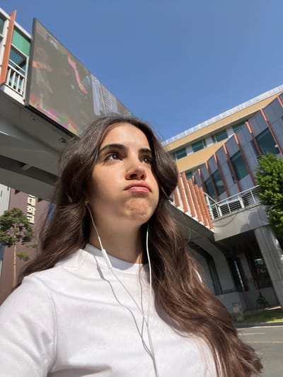 A low-angle, medium shot depicts a woman with brown hair wearing a white t-shirt and white earbuds, looking upwards. A building with a glass and metal facade is visible behind her. The sky is clear and blue. A sign with text in Korean Hangul is partially visible in the lower left corner of the frame, indicating a location in South Korea. Based on the script and architectural style, it can be inferred that the location is within an urban setting, likely in the city of Daegu, South Korea. The scene suggests an outdoor environment during daylight hours.