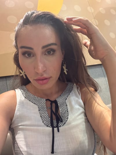 The media file is a selfie taken by a young woman in Nicosia, Cyprus. She has long, dark brown hair and is wearing a white top with black trim. She is also wearing gold starfish earrings. Her makeup is noticeable, with winged eyeliner and pink lipstick. She is touching her hair with her left hand. The background appears to be outdoors, with some large, pale circles on a textured surface. The lighting suggests it is daytime, possibly late afternoon, with sunlight casting shadows. The woman's expression is neutral, and the overall mood is casual. No visible text is present in the image.