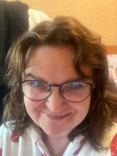 The image is a close-up selfie of a woman wearing glasses and a white and red patterned robe. She has brown, wavy hair and is smiling. The background appears to be a wall with a textured, yellowish-orange wallpaper. There are also hints of dark fabric and a white surface in the upper left corner, possibly clothing or bedding. No specific location in Tula, Russia can be identified from the image.