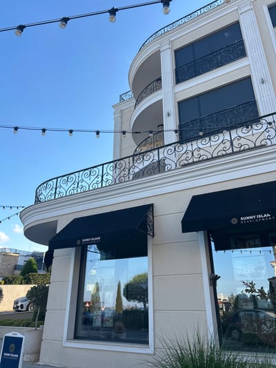 This media file captures a low-angle shot of a building facade under a clear blue sky. The building features classical architectural elements, including fluted columns and decorative wrought-iron railings on a balcony. String lights with exposed bulbs hang across the frame, suggesting an outdoor setting, possibly a commercial or residential area.
The facade includes large, dark-tinted windows, one of which displays an awning with the text "SUNNY ISLAND DEVELOPMENT" and a circular logo. Reflections in the window reveal the surrounding environment, including trees, other buildings, and what appears to be a parked car. The overall scene suggests it is daytime, with bright sunlight, and the weather is clear and fair.
The low angle of the shot emphasizes the grandeur of the building and the expanse of the sky. The composition creates a sense of upward movement, drawing the viewer's eye towards the architectural details and the clear sky. The presence of the string lights adds a touch of warmth and festivity to the otherwise formal architecture.
The visible text "SUNNY ISLAND DEVELOPMENT" indicates that this is likely a commercial property or part of a development project. The location context provided is Chernomorets, Bulgaria, which suggests a coastal or resort-like environment, possibly with a Mediterranean influence given the architectural style. There are no discernible people in the foreground, but the reflections in the window hint at activity within or near the building. The overall mood is serene and aspirational, showcasing the building's design and its integration with the environment.