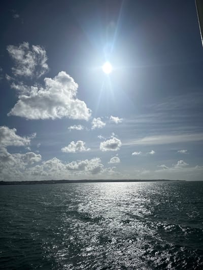 The image depicts a seascape with a bright sun in a partly cloudy sky over a body of water. The sun is positioned in the upper portion of the frame, emitting strong rays that create a glinting effect on the water's surface. Scattered white clouds drift across a blue sky. In the distance, a dark, elongated landmass with some buildings is visible along the horizon. The water is dark and choppy, reflecting the sunlight in a shimmering pattern. This scene is likely located near the city of Pointe-à-Pitre, France, given the context. There are no people or specific actions visible in the image.