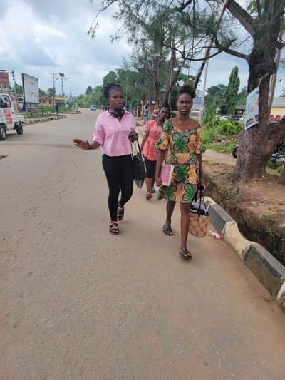 Three young women are walking on a paved road in Egbagu, Nigeria. The woman in the foreground is wearing a short, brightly colored green and orange patterned dress and black sandals. She is carrying a patterned tote bag and a pink mobile phone. The woman in the middle is wearing a pink short-sleeved shirt, black pants, and black sandals with pink accents. She has headphones around her neck and is carrying a black tote bag. The woman in the background is wearing a light orange, short-sleeved dress. The road is a light brown asphalt with a concrete curb and a shallow drainage ditch on the right side of the image. Trees line the right side of the road, with foliage and some taller trees in the background. The sky is overcast. In the background, there are buildings, streetlights, utility poles, and vehicles, including a white truck on the left.