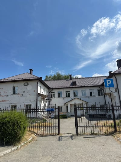 The image shows the exterior of a white, two-story building with a dark metal roof. The building appears to be an older structure, as evidenced by peeling paint on the exterior walls. There is a black metal fence in front of the building with an open gate. A blue sign with a white "P" and a wheelchair symbol is visible on the right side of the image. There is also a blue directional sign with Russian text that reads "Флюорография в здании стационара | 120" which translates to "Fluorography in the hospital building | 120". The sky above is blue with scattered white clouds. The scene is likely a medical facility. Based on the signage and typical architecture, this location is likely the building of the Altai Regional Clinical Hospital named after S.V. Zapolsky in Biysk, Russia, specifically the building housing the fluorography department.