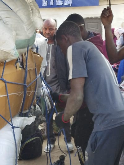 In Unguwa Uku Kauyen Alu, Nigeria, an outdoor scene depicts three males amidst cargo under bright daylight. A prominent stack of white woven sacks and cardboard boxes, secured by a blue rope net, occupies the left foreground. A young Black male in the center-right is seen from the chest down, wearing a dark blue t-shirt with a white vertical stripe and dark pants with "R AGRAMER" visible on the left leg. His hands, gloved in black with red trim, are actively engaged with the blue ropes, appearing dusty. Behind him, a second young Black male is partially visible, clad in a maroon hooded garment, with his right hand raised. Further back, a bald Black male with a beard and a dark spot on his forehead gazes towards the viewer, dressed in a grey traditional tunic with a white buttoned placket. The background features a light-colored building with a blue sign displaying white text that reads "Eat & Take Away." The ground is unpaved, appearing as dirt or sand.