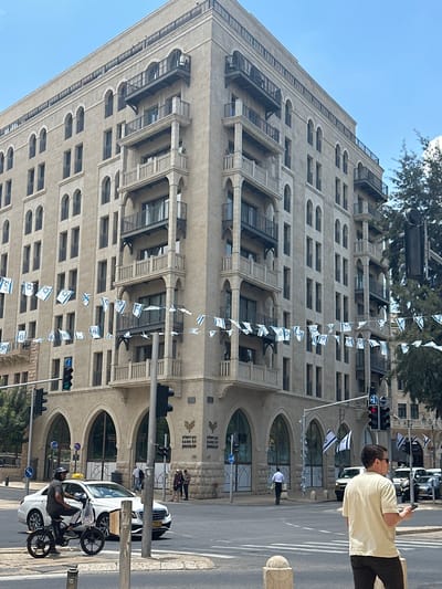 A street scene in Jerusalem, Israel, shows a multi-story building with light-colored stone or concrete facade. The architecture includes repeated patterns of windows and arched details. Strings of Israeli flags hang vertically along the building's front. A man in a light-colored shirt is standing on the sidewalk looking at his phone. Street-level includes sidewalks, traffic lights, and traffic signs. Cars are parked along the side of the road and a man is riding a motorcycle. A sign on the building identifies it as "Bank of Jerusalem." Pedestrians are walking near the entrance of the bank.