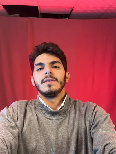 A young man with dark hair and a neatly trimmed beard is shown from the chest up. He's wearing a gray sweater over a white collared shirt. The background is a gradient of red, lighter in the middle and darker on the edges. To the left, there is a partially visible television mounted on the wall. The man looks directly at the viewer with a slight tilt of his head.