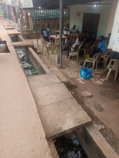 The image depicts an outdoor scene in what appears to be a neighborhood in Enugu, Nigeria, characterized by a visible drainage channel running alongside a paved walkway. Several individuals are seated at tables, suggesting a communal gathering or a local eatery. The area seems to be somewhat utilitarian, with a mix of seating, tables, and a blue plastic crate, possibly used for beverages. The presence of a drainage channel with visible debris indicates an urban environment where infrastructure management might be a concern. The lighting suggests it is daytime.