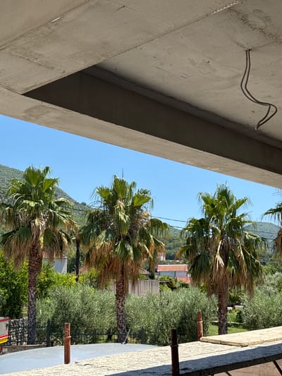 The image depicts an outdoor scene with a clear blue sky. In the foreground, a concrete structure with exposed rebar and electrical wires extends across the top of the frame. Below this structure, a row of palm trees with lush green fronds and dry lower trunks are visible. Behind the palm trees, there is dense green foliage, possibly olive trees, and some buildings with red roofs. In the background, rolling green hills dominate the landscape. The scene is situated in Bratešići, Montenegro.