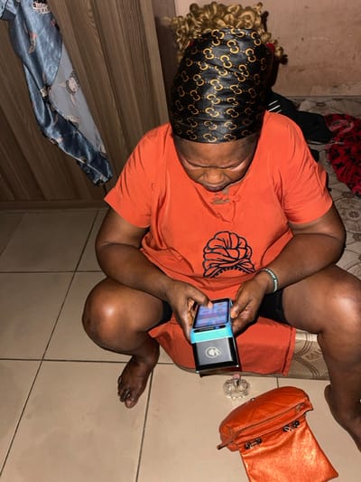 An individual wearing an orange shirt and a patterned headscarf is seated, legs crossed, on a tiled surface. They are holding a black and teal device, seemingly focused on its screen. Nearby is an orange purse and a transparent object, possibly a bottle or decorative piece. The setting appears to be indoors, indicated by the presence of furniture and a wall with visible wear. Other articles of clothing are seen in the periphery.