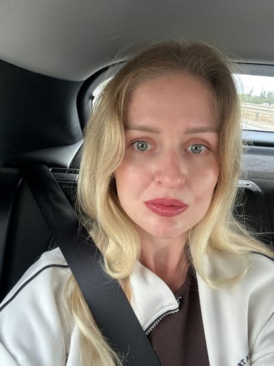 A woman with blonde hair and green eyes is in a car wearing a seatbelt and a white jacket with black trim. The interior of the car is black. The location is likely Petah Tikva, Israel, based on the request, but there are no visible details in the image confirming this.