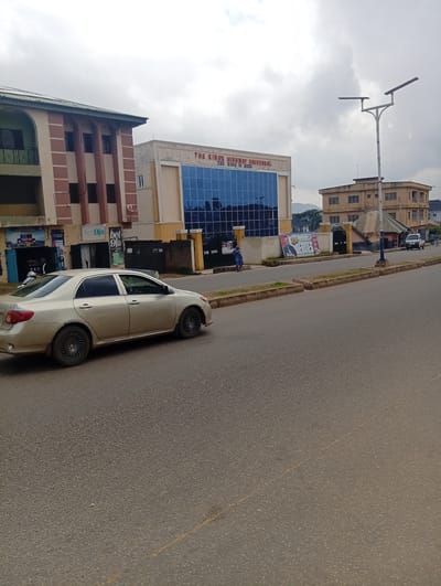 A daytime, eye-level, outdoor, medium shot captures a street scene in Kabong, Nigeria. In the foreground, a silver Toyota Corolla sedan is parked on the right side of a paved road. The car is facing left, with its rear end closest to the viewer. The road itself is asphalt with faint yellow lane markings visible. A sidewalk with grass and concrete curbing runs along the right side of the road.
In the midground, to the left, is a multi-story building with a tan and red facade. It has arched windows and appears to be a commercial establishment. Next to this building, to the right, is a modern, two-story building with a light-colored exterior and a large glass facade on the upper level, reflecting a blue sky. Red lettering on this building reads "THE KING'S HIGHWAY UNIVERSAL THE KING IS HERE". To the right of this building, a street lamp with two horizontal fixtures stands tall. Further to the right, a three-story yellow building with multiple windows is visible.
In the background, a hazy, mountainous landscape can be seen. The sky is overcast with a mix of gray clouds. Several people are visible: a woman in a blue dress stands on the sidewalk in front of the modern building. A man in dark clothing stands inside a doorway of the tan and red building. A few other figures are sparsely distributed along the street. A white pickup truck is parked near the yellow building. Various signs are posted on the buildings, including advertisements for "bet9ja".