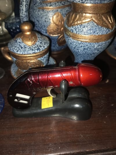 A small, red, novelty gun sits on a black stand. The gun, metallic red with black handle and trigger, is positioned in the center, resting on its stand. Behind the gun are various decorative ceramic pieces: a small, lidded pot and two vases, all painted with a blue and gold pattern. They add a patterned backdrop to the scene, which is set on a dark wooden surface.