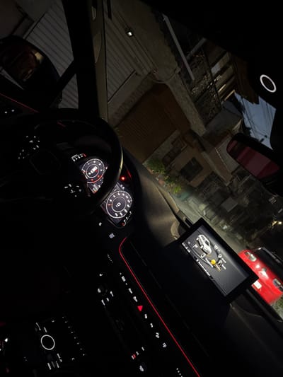 The image shows the interior of a car at night, with the dashboard and infotainment screen illuminated. The setting appears to be a residential street, with buildings and a red car visible through the windshield. The infotainment screen displays the car's menu, with options for "Car," "Sound," "Radio," "Media," and "Telephone." The instrument cluster shows the speedometer, tachometer, and other driving information. The time is displayed as 5:15. The overall mood is one of quiet introspection, perhaps a moment of rest or contemplation before continuing a journey. The visible text includes "Sound," "Radio," "Media," "Telephone," "Bluetooth audio player," "MAX," "AUTO," "REAR," "MENU," "BACK," and "MEDIA," along with numbers on a keypad and a mileage reading of "231251." The location context is Gotse Delchev, Bulgaria.