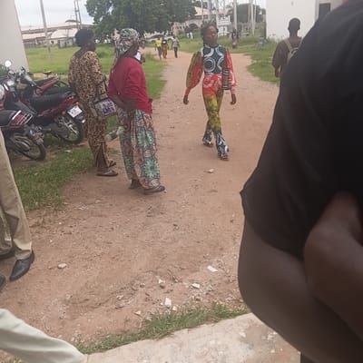 In Wukari, Nigeria, several individuals are gathered outdoors on a dirt path adjacent to a grassy area. The scene appears to be during the daytime, under overcast skies.
The central figures are three women dressed in vibrantly patterned traditional African attire. One woman, in the foreground and to the right, wears a red top and a long skirt with a colorful, intricate floral design. Another woman, to her left, is clad in a brown and white patterned dress and a green headscarf. A third woman, further back and to the left, is dressed in a striking outfit featuring a red, white, and green patterned top with puffy sleeves and a matching green and yellow patterned skirt. Her eyes are closed, and her arms are slightly outstretched. Several other people are visible in the background, appearing as smaller figures walking or standing.
Two motorcycles are parked on the dirt path, one red and one darker in color, with visible license plates. One of the license plates appears to have the letters "DD" and "134" visible. A portion of a white building with windows is visible in the upper left of the image, along with some infrastructure that could be electrical poles or similar utility structures. The ground is a mix of dirt and small stones, with patches of green grass along the right edge of the frame. The overall atmosphere seems to be one of a casual gathering or community event.