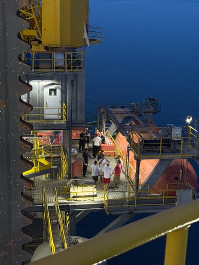 In the image, a group of people are gathered on a metallic platform of an industrial structure, possibly an oil rig or a similar offshore facility. The structure is characterized by its yellow and gray metallic components, safety railings, and stairs. One section of the structure features a large, orange capsule, possibly a life raft or escape pod, mounted on a mechanism. The people are dressed in casual clothing, and their interactions are not clearly discernible from the image. The background is a vast expanse of dark blue water, suggesting an offshore or maritime setting. The lighting indicates it is either dusk or night. The specific location in the city of Unknown, Italy, cannot be determined from the provided image.
