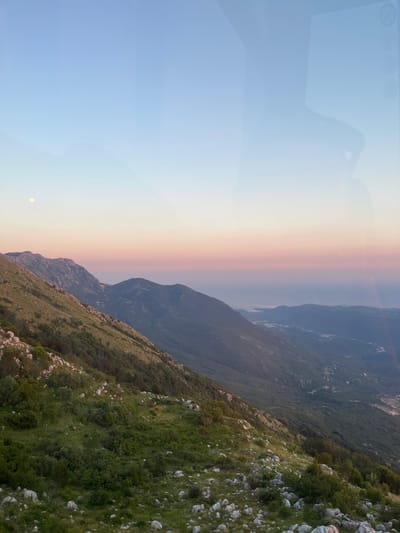 A high-angle, wide shot captures a mountain landscape at dusk. The sky transitions from a pale blue at the top to soft pinks and oranges near the horizon, where a faint, pale moon is visible in the upper left quadrant. The foreground features a steep, grassy mountainside dotted with white rocks and scattered bushes. The midground consists of rolling, tree-covered hills, and in the distance, a hazy body of water merges with the sky. A subtle reflection of what appears to be a window frame or a similar structure is visible on the left side of the image, overlaying the sky and the distant landscape. The scene depicts a serene, natural environment with a sense of vastness.