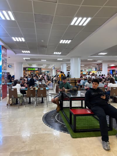 This is a candid, vertical shot of a busy food court, likely in an airport or large shopping mall, given the array of restaurants and seating areas. The scene is brightly lit by overhead fluorescent lights, suggesting it's daytime. The floor is tiled with a marble pattern.

The primary subjects are the many people seated at tables, engaged in eating, talking, or waiting. The crowd is diverse in age and ethnicity. In the foreground, a person is seated on a bench with a red top and black metal frame, set on a patch of artificial green turf that resembles a small soccer field. This person is wearing dark clothing and grey Croc-like shoes. Another person, wearing a green t-shirt and plaid shorts, is seated at a table and looking towards the camera, seemingly in conversation.

Visible text includes "STAR ISLAND CAFÉ" on a sign above one of the food stalls. Other signs with text like "SETE NTOJA?" and "CANG SOMETHING" are also present, along with what appears to be menu boards and advertisements for food. The style of the food court and the presence of multiple eateries suggest a public space designed for dining and relaxation. The atmosphere appears lively and somewhat chaotic due to the number of people present. The mention of "Cancún, Mexico" as location context suggests this could be an airport food court in Cancún.