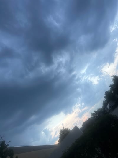 The image shows a low-angle view of a cloudy sky, possibly taken during sunset or sunrise, in Schweitenkirchen, Germany. The sky is dominated by large, dark clouds in the foreground, suggesting an approaching storm or the aftermath of one. In the upper right portion of the image, the clouds lighten and break, revealing patches of a brighter sky, tinged with a hint of orange, characteristic of twilight. Silhouetted against the sky are the tops of dark green trees and the edge of a light-colored building, likely a multi-story structure. A street or road is visible at the very top edge, with what appears to be a white vehicle. The overall mood is dramatic and somewhat somber due to the heavy cloud cover, but the subtle colors in the distance offer a touch of hope. No text is visible in the image.