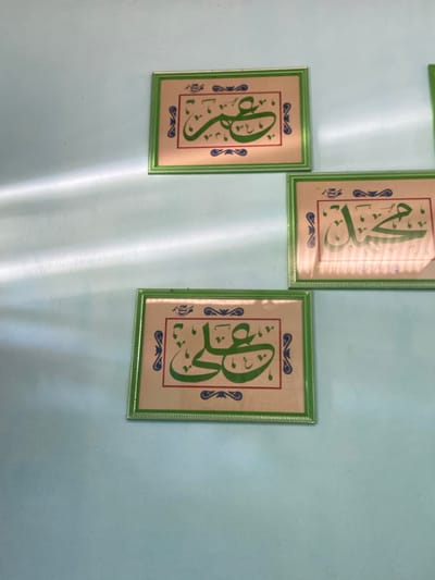 Three framed pieces of Islamic calligraphy are displayed on a light blue wall. The frames are green with decorative borders. The calligraphy is in green ink on a light background, with blue decorative elements. The arrangement suggests a decorative display, possibly in a home or public space within Sekar Mawar, Indonesia. The lighting on the wall indicates the presence of light sources creating shadows and highlights, suggesting daytime.