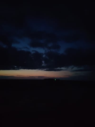 A dark, grainy, landscape photograph depicts a twilight scene in Larionovo, Russia. The sky is filled with ominous, dark clouds, obscuring most of the celestial bodies. A sliver of orange and pink light peeks through the horizon, indicating the last vestiges of daylight. The foreground is a silhouette of the darkened land, with a faint, distant structure visible against the horizon, marked by two small green lights.