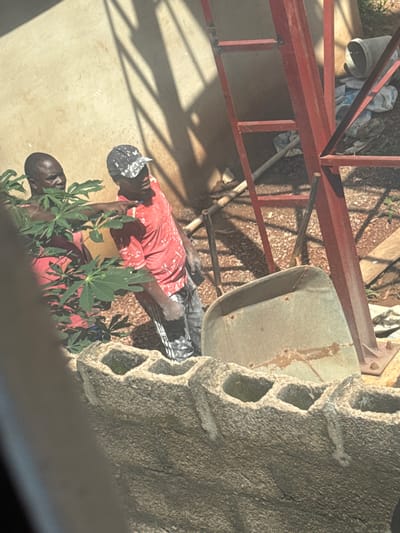 Two men are working outdoors. The man on the left is wearing a red t-shirt and is partially obscured by green foliage. The man on the right is wearing a red t-shirt with white paint splatters and a black and white patterned baseball cap. He is pointing to the left with his right hand. Behind them is a concrete wall, a red metal ladder structure, a wheelbarrow, and scattered building materials. The overall scene suggests a construction or renovation project. The presence of a red metal ladder structure and building materials suggests this scene is located in Enugu, Nigeria, likely a residential or commercial construction site.