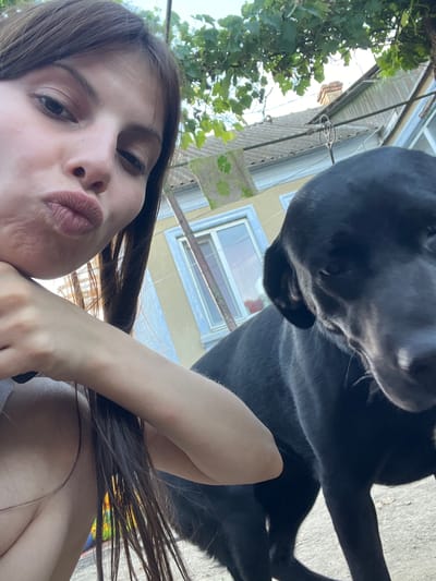 A low-angle, close-up selfie taken outdoors shows a young woman with long, dark brown hair and a fair complexion. She is puckering her lips and resting her chin on her hand, with her left arm visible. Next to her, and partially obscuring the background, is a black dog with its head facing the camera, appearing to be looking down. The background consists of a building with yellow walls and a light blue trim around a window. Above the building, a grey, tiled roof is visible, with green foliage hanging from trees and vines. The lighting suggests it is daytime, possibly late afternoon or early evening, with sunlight casting shadows. The scene is set in an outdoor environment, likely a yard or courtyard, with a concrete ground surface. No specific landmarks in Blizhnii Khutor, Moldova are identifiable.