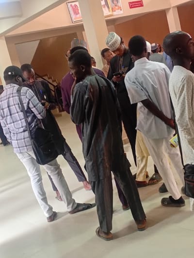 The image displays an indoor scene in the city of Kwazailewa, Nigeria, populated by approximately ten to fifteen men. The setting is a brightly lit hall or lobby with a smooth, light-colored floor and neutral-toned walls. In the upper background, two framed portraits and a small red rectangular sign are mounted on a wall. A dark-colored staircase with a visible handrail ascends on the left side of the background.
The individuals are primarily young to middle-aged adult males, dressed in a combination of traditional Nigerian garments, such as kaftans and agbadas, and casual attire including shirts and trousers. Several men wear traditional caps.
In the immediate foreground on the left, a man wearing a light plaid shirt, light trousers, and a dark cap carries a black shoulder bag and is focused on a smartphone. Adjacent to him, another man, seen from behind, wears a dark, possibly striped, traditional two-piece outfit and open-toe sandals. To the right of the foreground, a man in a dark traditional outfit and a white/grey patterned cap is looking down at a smartphone. Further back, another man is seen from behind, wearing a light blue/grey short-sleeved shirt and light trousers. Most individuals are standing, with several observed examining their smartphones. No direct physical interactions or explicit conversations are discernible.