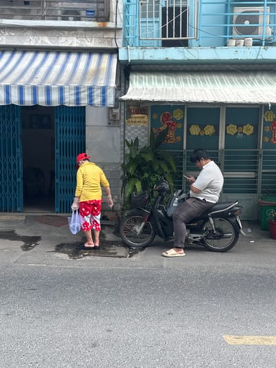 The image displays an urban street scene in Ho Chi Minh City, Vietnam. The foreground shows an asphalt road with a faded yellow line marking. The midground features a concrete sidewalk with several puddles.
On the left, an individual, likely female, is walking towards an open blue accordion-style metal security gate. She wears a red baseball cap, a yellow long-sleeved shirt, red and white patterned capri pants, clear plastic gloves on both hands, and black slip-on sandals. She carries a clear plastic bag in her left hand. The building behind her is light grey with a blue and white striped retractable awning extended over the entrance. An "AQUA" air conditioning unit is visible on a higher balcony.
To the right, a male individual is seated on a black motorbike parked at the edge of the sidewalk. He wears a white and grey striped polo shirt, dark grey pants, and light beige slip-on sandals. He is looking down intently at a smartphone held in both hands. A dark grey backpack rests on the motorbike's seat behind him. The building behind him is light blue with a faded blue and white striped awning. Its ground floor features a decorative gate with red horse and gold flower motifs. A large green plant stands between the two buildings. A green plastic trash receptacle is partially visible on the far right. An "TOSHIBA" air conditioning unit is mounted on an upper balcony of the blue building. Various signs with Vietnamese text are affixed to both structures.