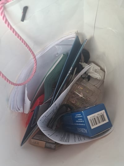 This is an eye-level, indoor, medium shot of a white paper shopping bag filled with various items. The bag's left side has a pink braided rope handle. Inside the bag, from left to right, are several stacks of papers, some appearing to be documents or notebooks. Further in, there is a dark blue folder or binder. Next to it, a white USB wall charger with the brand "Infinix" is visible. To the right of the charger is a clear, rectangular plastic cup adorned with colorful cartoon drawings of houses, flowers, stars, and unicorns. Beneath the cup and charger, a small, clear glass bottle with a white lid is partially obscured. In the lower right of the bag is a blue cardboard box with the text "AL'DENTE" and a barcode. In the lower left, a pack of cigarettes with the brand "LEX" is visible. The background is the white interior of the shopping bag. The scene appears to be set in Leningradskaya, Russia, given the Armenian script on some of the documents, which suggests a connection to the Armenian community or businesses in the region, and the presence of the USB charger which could be used in that city.
