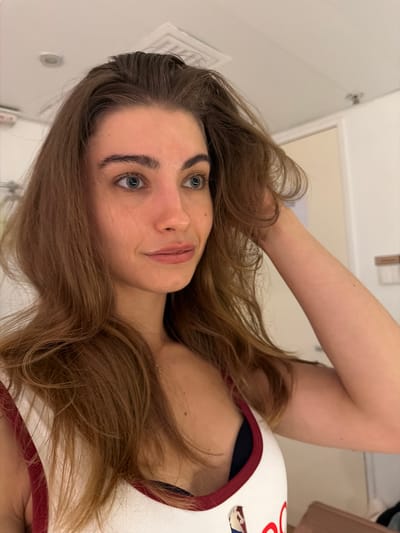 This is a medium shot, portrait-style photograph of a young woman with long, light brown hair. She is looking to her right, with her left hand touching her hair. She is wearing a white tank top with red trim, and a dark bra is visible underneath. The background appears to be an indoor setting, possibly a hotel room or dressing room, with a white ceiling, a light-colored wall, and a door in the background. There is a vent or light fixture on the ceiling. The image does not provide any specific information or visual cues to indicate a location in Hong Kong, China.