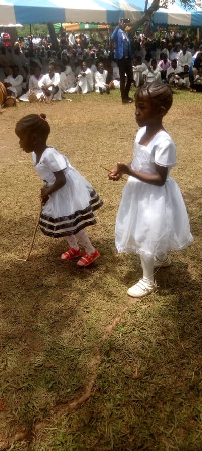 Two young girls are standing in a field, wearing white dresses. The girl on the left is holding a stick, while the girl on the right is holding a stick. The ground is covered in dry grass. There is a large group of people sitting in the background, under a white and blue striped tent. The event is being held in Burundu, Kenya, a suburb of Nairobi. The coordinates provided (latitude: 0.46, longitude: 34.776) correspond to a location within the suburb of Burundu, Kenya.