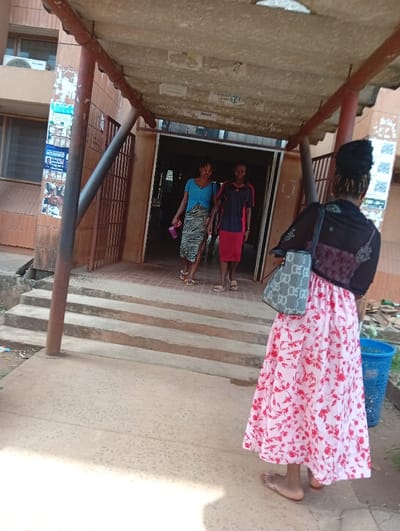 A daylight outdoor scene in Nsukka, Nigeria, depicts three individuals near a building entrance with concrete steps and an overhead canopy. In the foreground, one individual, seen from behind, wears a black top, a long pink skirt with red floral patterns, and light-colored sandals, carrying a patterned shoulder bag on her left side. She faces a building entrance. From this entrance, two individuals walk down three concrete steps towards the foreground. The person on the left wears a light blue top, a patterned skirt, and light-colored sandals, holding a pink bottle. The person on the right wears a dark polo shirt with red accents, a red skirt, and light-colored sandals. The building's left wall features brown tiling, a window, an air conditioning unit, and various posters. The underside of the overhead canopy is discolored and has posters affixed, showing partial text like 'CHRIST' and 'ISAAC'. A blue plastic bin is visible to the right of the steps. The ground is a light-colored concrete slab.