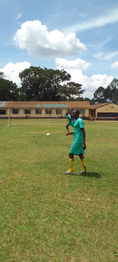 The image shows a man wearing a black face mask and a teal shirt and yellow socks walking on a grass field in Lubao, Kenya (latitude: 0.384, longitude: 34.801). There is a soccer goal in the background, and a soccer ball is lying on the grass. The man is looking towards the left of the image, and the sky is blue with white clouds.  There is a building behind the soccer goal, which appears to be made of concrete, with windows and a brown roof.  To the right of the building, a tree line can be seen. In the background, other people can be seen. There is a brown, grassy field to the right of the man.  The grass field is mostly covered in green grass, with some patches of brown.