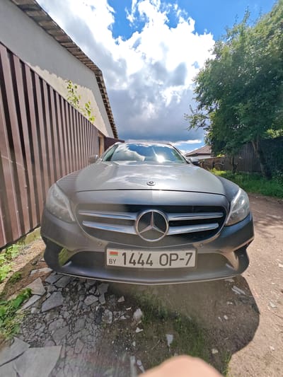In a frontal, low-angle shot, a dark gray Mercedes-Benz sedan is parked on a gravel and cracked asphalt surface. The car faces forward, with its headlights illuminated. The license plate, visible on the front bumper, reads "1444 OP-7" and features the Belarusian flag. To the left of the car is a tall, dark brown metal fence adjacent to a light-colored building with a weathered roof. To the right, a lush green tree with dense foliage extends over the car. In the background, a rustic wooden fence and a residential building with a red-tiled roof are visible under a bright sky filled with dramatic white and gray clouds. The scene appears to be in a rural or suburban area. The lighting suggests it is daytime, possibly late morning or early afternoon, given the shadows cast.