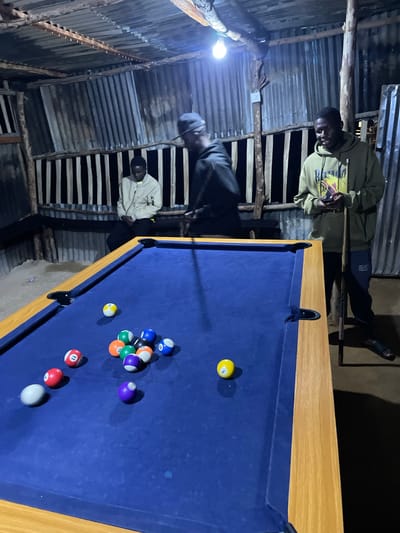 In a rustic, dimly lit structure in Wumat, Nigeria, three individuals are engaged in a game of pool. The walls are constructed from corrugated metal sheeting and wooden planks, suggesting a simple, possibly makeshift, establishment. A bare light bulb hangs from the ceiling, casting a stark illumination on the scene.
The central focus is a blue pool table with a wooden frame. Several pool balls are scattered across the table. Visible balls include a yellow ball with the number "1," an orange ball with "3," a green ball with "9," a red ball with "11," a purple ball, a white cue ball, a blue ball with "10," a black ball with "8," and a blue and white striped ball with "12." The arrangement of the balls suggests the game is in progress.
Two men are actively involved in the game. One man, wearing a light green hoodie and dark pants, is holding a pool cue and appears to be taking a shot. He has a focused expression. Another man, in a light-colored long-sleeved shirt and dark pants, is positioned near the table, observing. A third individual, partially visible in the background, is also present.
The time of day appears to be evening or night due to the artificial lighting and dark surroundings. The overall atmosphere is casual and social, with the individuals engrossed in their game. The rough construction of the building and the simple lighting contribute to a raw, authentic feel of the location. No specific weather conditions are discernible. There is text on the green hoodie of the man taking the shot, but it is not clearly legible. There is also text on the pool cue.