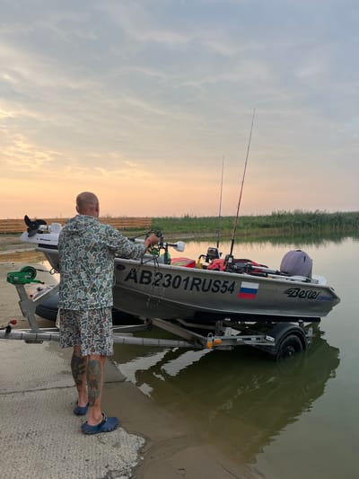 A bald man in camouflage clothing stands next to a boat on a trailer at the edge of a body of water. The boat is silver with the license plate "AB2301RUS54" and a Russian flag sticker on its side. Fishing rods are placed in the boat, and a trolling motor is attached to the stern. The man appears to be preparing the boat, holding ropes in his hands. The environment consists of a sandy shore, concrete ramp, and lush green vegetation in the background. The sky is overcast with hints of pink and orange from a sunset. The trailer's wheels are partially submerged in the water, suggesting the boat is being launched or retrieved. The image appears to be taken in Zav'yalovo, Russia, given the license plate.