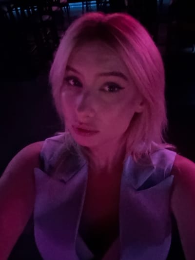 The image is a close-up, selfie-style photograph of a young woman with blonde hair, illuminated by pink and purple lighting, suggesting a dimly lit indoor environment. She is wearing a light blue vest. The background is dark and indistinct, with hints of blurry shapes that could be furniture or other patrons, indicating a social setting like a bar or club. There is no visible indication of the city of Amman, Jordan, or any specific geographic markers.