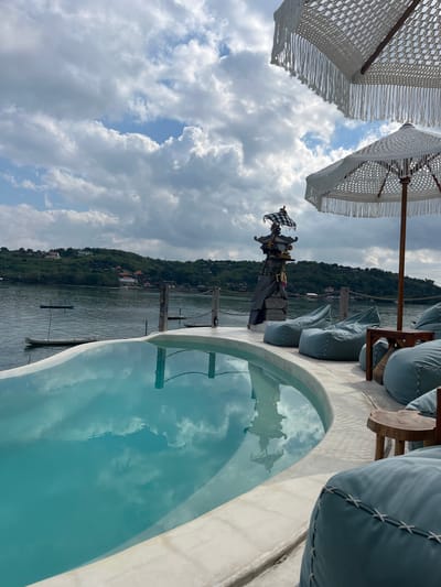 This is a medium shot, taken from a slightly high angle, of an infinity pool overlooking a body of water. The pool is in the foreground, its turquoise water reflecting the cloudy sky. To the right of the pool, on a white concrete deck, are several light blue beanbag chairs and a small wooden table. In the background, across the water, is a green, hilly coastline with scattered buildings. Several small boats are visible on the water. Two white umbrellas with decorative fringe are partially visible at the top of the frame, suggesting an outdoor seating area or resort. The scene appears to be in Nusa Penida, Indonesia, given the distinctive landscape and the Balinese-style altar-like structure visible behind the pool on the left.