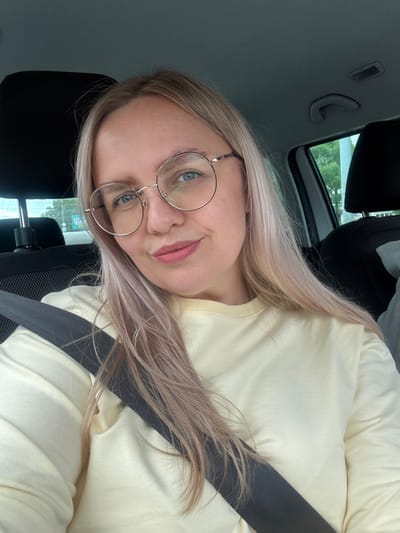 The media file shows a young woman with blonde hair, wearing glasses and a light yellow sweatshirt, seated in a car. The image is taken from a close-up perspective, focusing on her face and upper body. She is looking at the camera with a slight smile. A black seatbelt is visible across her chest. The background of the image shows the interior of the car, including a car seat, and through the window, greenery and trees can be seen, suggesting an outdoor setting. The lighting indicates it is daytime. There is no visible text in the image. The setting appears to be Vologda, Russia, based on the provided location context.