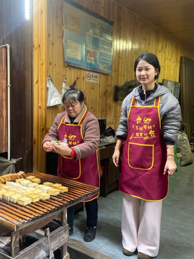 Two women wear matching maroon aprons with gold trim and Chinese characters. The younger woman stands smiling, wearing a gray puffer jacket and white pants. The older woman, in a brown sweater and dark pants, focuses on packaging small items. They stand inside a room with wooden walls and floors. A display board with documents and a "No Smoking" symbol is visible on the wall. Rows of small, square objects are displayed on a wooden rack.