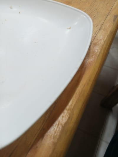 The image shows a white square plate on a wooden table. There are a few crumbs on the plate. The lighting is dim, and the focus is on the plate and the table. The background is blurred and appears to be a tiled wall and a dark shadow. The scene is likely from a domestic setting.