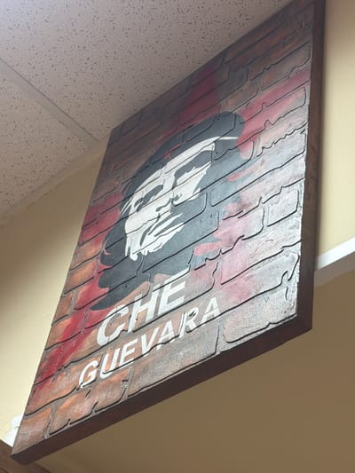 This media file shows a close-up, angled view of a textured artwork. The artwork appears to be a mural or a sign depicting a portrait and text. The textured surface mimics brickwork, with raised lines forming rectangular shapes that suggest mortar lines. The colors used are predominantly muted reds, browns, and grays, with stark white and black for the portrait and text.
The central subject of the artwork is a stylized black and white portrait, seemingly of a person with distinct facial features. To the right of the portrait, white text is prominently displayed. The visible text reads "CHE GUEVARA". The artwork has a rustic, possibly distressed aesthetic.
The setting appears to be indoors, as suggested by the textured ceiling tiles visible in the lower left corner of the image and the plain wall to the right. The lighting is neutral, not indicative of any specific time of day. There are no clear environmental cues to suggest the location is Maloyaroslavets, Russia, other than the provided metadata. The artwork itself does not contain any specific Russian cultural elements. The overall impression is one of a decorative piece, perhaps a mural in a public space or a decorative sign.