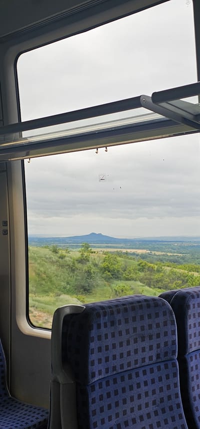 The image captures a view from a train window, overlooking a scenic landscape. In the foreground, the dark blue, patterned upholstery of train seats is visible. The window reveals a rolling green hill dotted with trees and shrubs. In the distance, a prominent mountain with a distinct peak rises against a cloudy sky. The sky itself is overcast with a diffused light. The train's interior structure, including a metal bar and window mechanisms, is also present. On the left side of the window, a label with the numbers "105" and "107" is visible. The scene is devoid of people. The location is identified as Kozar Belene, Bulgaria.