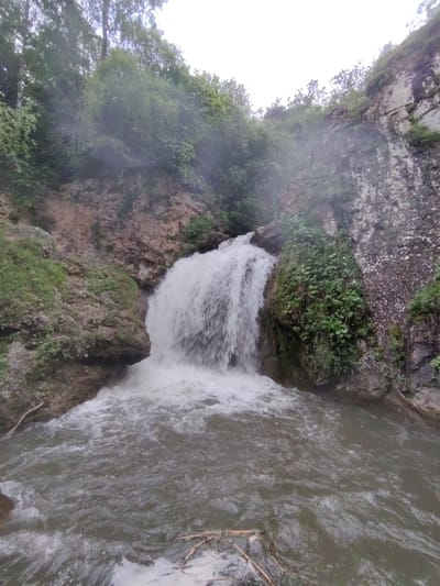 This media file shows a waterfall in a natural setting. The waterfall is the main subject, with water cascading down a rocky cliff face into a pool below. The surrounding area is lush with greenery, including trees and bushes clinging to the rocky slopes. The overall scene is one of natural beauty and tranquility.

The time of day appears to be overcast, likely daytime, due to the diffused light and lack of harsh shadows. The weather seems to be humid or possibly rainy, as there are water droplets visible on the lens, obscuring some details and giving a misty appearance to the scene. The location is identified as Krasny Kurgan, Russia.

There are no people or man-made objects clearly visible in the scene, emphasizing the raw, untouched nature of the environment. The activity taking place is the natural flow of water forming the waterfall. The emotion evoked by the image is one of serenity and the power of nature. There is no visible text in the media file.