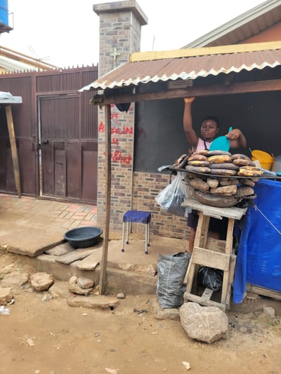 A person is standing behind a stall, holding a blue watering can and a bunch of plantains. The stall is covered by a corrugated metal roof and has a brick wall in the background. There are more plantains and some rocks displayed on the stall. To the right of the stall, there is a wooden structure with a blue tarp draped over it, and a black garbage bag inside. The ground is a mix of dirt and concrete. In the foreground, there is a black basin and some scattered stones. The scene appears to be outdoors, possibly in a market setting in Egbagu, Nigeria. The lighting suggests it is daytime. There is some red graffiti on the brick wall behind the stall.